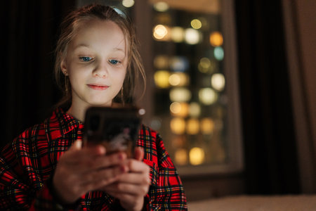 Portrait of pretty little girl browsing smartphone screen, nighttime cityscape reflecting on window, soft digital glow illuminating happy facial expression, enjoying mobile apps, games, social media.の写真素材