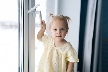 Portrait of curious adorable child girl holding touching toward window handle, looking at camera. Concept of potential home safety risks for curious preschoolers exploring indoor spaces.の写真素材