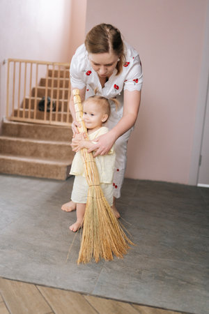 Caring mom teaching baby girl how to sweep floor with broom, promoting early learning and household chores in playful and educational way. Concept on developing self-care habits, childhood routine.の写真素材