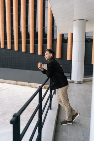 Vertical side view shot of pensive man enjoying coffee break, leaning against railing outside modern office building, thoughtful looking away at cityscape. Concept of modern successful city lifestyle.の写真素材
