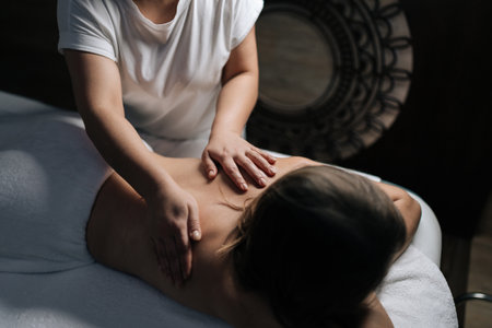 Close-up of professional female masseuse giving relaxing back massage to young unrecognizable woman lying on massage table in spa salon with dark interior, creating tranquil and soothing atmosphere.の写真素材