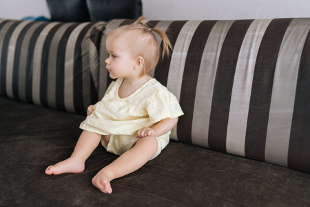 Portrait of blond-haired toddler wearing pastel yellow dress, relaxing sitting on striped couch, looking sideways with innocent curiosity in soft-lit living space. Concept of pleasures childhood.の写真素材
