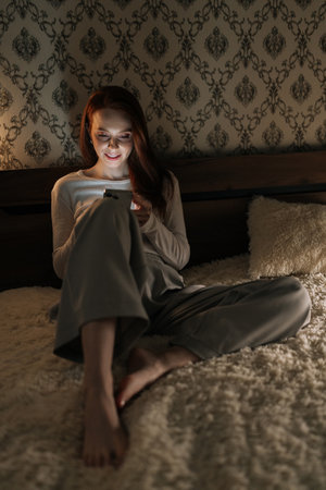 Vertical portrait of cheerful woman relaxing in dark bedroom, scrolling social media on smartphone,...