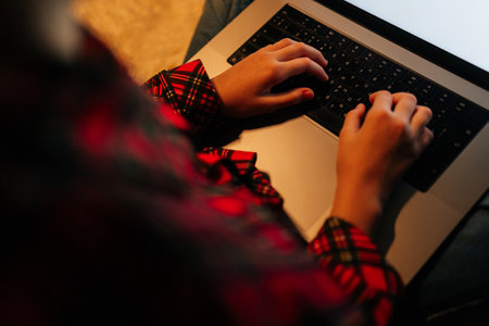 View from shoulder of unrecognizable child girl wearing red and black checkered shirt typing on backlit laptop keyboard sitting on bed with crossed legs in dark room, creating cozy atmosphere.の写真素材