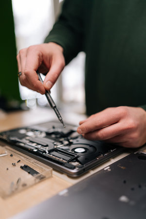 Vertical cropped shot of electronics repair professional carefully disassembling laptop motherboard, precision screwdriver in hand, surrounded by various components on workshop table, close-up.の写真素材