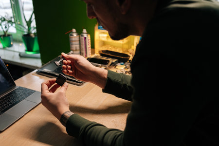 Focused technician repairing smartwatch battery using tweezers for precise component extraction and replacement in dark workshop sitting at desk with laptop computer.の写真素材