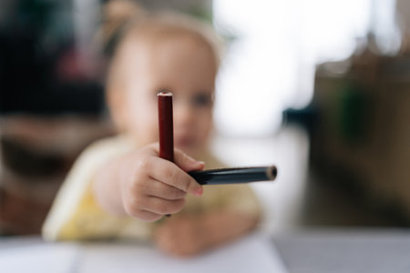 Blurred toddler girl showing two pencils to camera while sitting at table and drawing on paper at home, selective focus. Concept of childhood development, early learning, education.の写真素材
