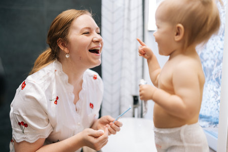 Mother helping toddler daughter brushing teeth together, playfully touching nose in bathroom, sharing joyful morning hygiene routine. Concept on developing self-care habits and early childhood routineの写真素材