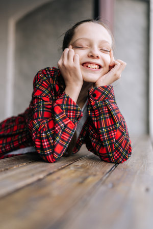 Joyful 10 years old girl with braided hair, wearing red checkered shirt, with eyes closed, smiling and holding face with hands, conveying sense of joy and happiness, leaning on rustic wooden table.の写真素材