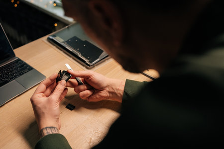 View from shoulder of professional technician carefully replacing smartwatch battery with precision tweezers, highlighting delicate electronic repair process, sitting at desk in dark workshop.の写真素材