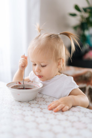 Vertical shot of toddler girl happily eating healthy soup with spoon, savoring meal at home, surrounded by warmth and love in cozy kitchen room. Concept on developing self-care habits.の写真素材
