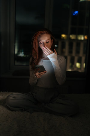 Vertical portrait of tired woman yawning and covering mouth with hand browsing internet, chatting in social media at night sitting by window with blurred bokeh city lights, in dark room.の写真素材