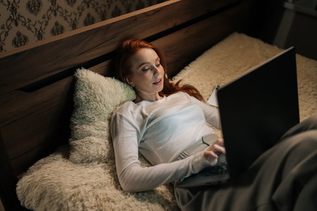 Top view of young female freelancer working typing on laptop with focused expression, lying comfortably on bed at night, smiling looking to screen, creating cozy and productive atmosphere in bedroom.の写真素材