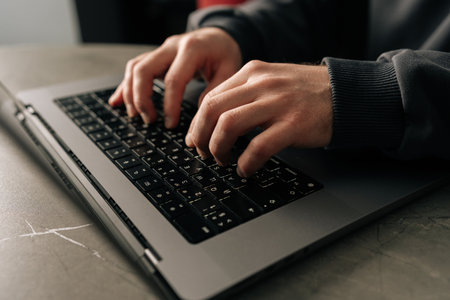 Close-up cropped shot of unrecognizable programmer male typing on laptop keyboard, immersed in software development or coding, sitting at table by window in dark room. Concept of remote job, freelanceの写真素材