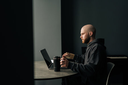 Side view of concentrated freelancer male with wrist brace working on laptop computer at home, enjoying coffee sitting in dark room. Concept of dedication and perseverance in remote work environment.の写真素材
