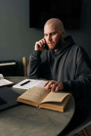 Vertical portrait of music composer talking on phone and reading book at home studio, with laptop and sheet music on table, working remotely on new project. Concept of remote job, technology, people.の写真素材