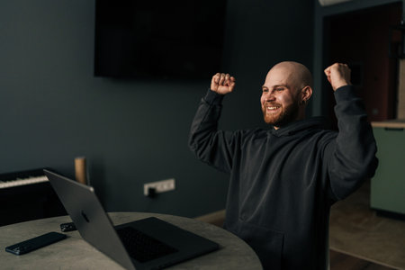 Cheerful male freelancer joyfully raising arms while working from home office, celebrating successful project completion with enthusiastic body language. Concept of remote job, technology and people.の写真素材