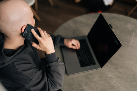 High-angle view of bald freelancer male working remotely, communicating on smartphone and typing on laptop, managing business while multitasking in contemporary home workspace, sitting at desk.の写真素材