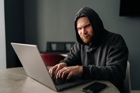 Portrait of funny male in black hoodie smiling looking at camera while typing on laptop keyboard.の写真素材