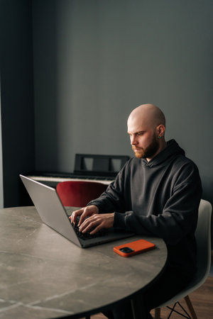 Vertical portrait of young bald programmer with beard and ear piercing sitting at round table in home office, concentrating on work on laptop computer looking to screen, mobile phone nearby.の写真素材