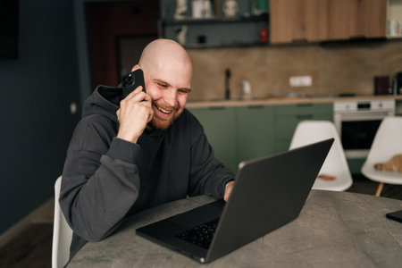 Happy smiling freelancer in hoodie remote working from home office, using laptop computer and talking on mobile phone, enjoying productive and comfortable work environment, sitting at kitchen table.の写真素材