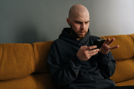 Close-up of serious young bald man with beard and hoodie using smartphone while sitting on comfortable sofa, browsing internet, social media or mobile application. Concept of modern technology.の写真素材