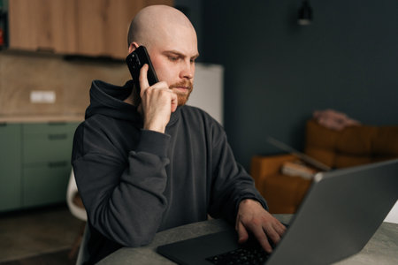 Serious freelancer male working from remote home office, talking on mobile phone and using laptop computer, managing business and multitasking in home environment. Concept of remote job, technology.の写真素材