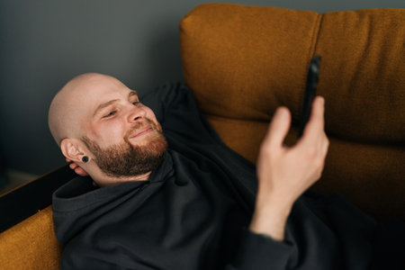 Close-up face of cheerful man with beard wearing dark hoodie lying comfortably on sofa using mobile phone, enjoying leisure time at home. Resting happy bald male surfing cellphone lying couch indoors.の写真素材