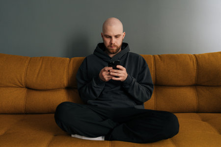Portrait of young man with beard in casual clothes, sitting cross-legged on comfortable sofa, using mobile phone while enjoying relaxing moment at home. Resting bald male surfing cellphone indoors.の写真素材