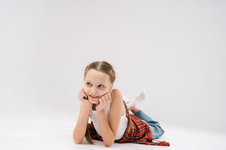 Adorable smiling young girl lying on floor with chin resting on hands, wearing white tank top and red checkered shirt, radiating playfulness and carefree joy. Concept of happy childhood.の写真素材