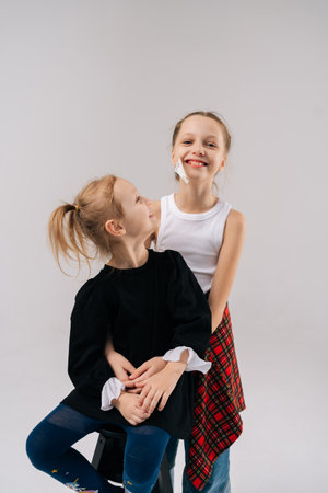 Loving sisters hugging closely in white studio setting, elder sibling standing behind younger one seated on stool, sharing warm emotional connection, genuine happiness together.の写真素材