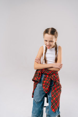Funny preteen girl with braided hair wearing jeans and red plaid shirt tied around waist, sticking out tongue and folding arms, conveying playful and mischievous attitude, smiling looking at camera.の写真素材