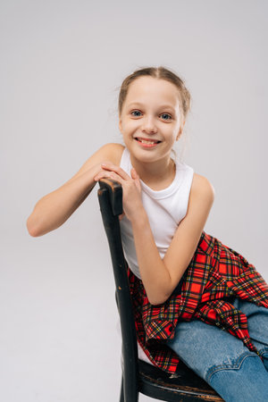 Vertical studio portrait of smiling preteen girl with blue eyes and light brown hair, wearing white tank top, red checkered shirt and jeans, sitting on vintage black chair on white isolated backgroundの写真素材