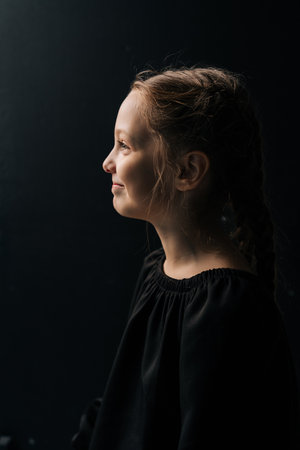 Side view studio portrait of smiling young preteen girl with braided hair, wearing black dress, posing against dark isolated background. Closeup face of pretty child illuminated by soft light.の写真素材