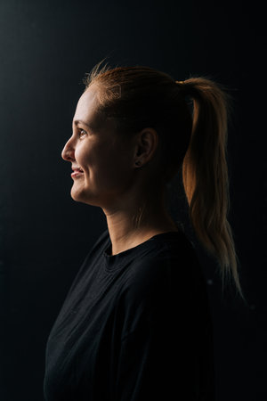 Side view vertical portrait of positive young woman with ponytail, wearing black t-shirt, smiling looking away against dark background in studio setting, close-up face illuminated by soft light.の写真素材