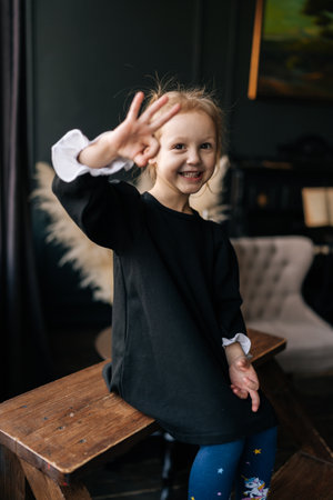 Cheerful child wearing black dress and colorful tights showing OK sign with fingers while sitting on wooden bench in cozy home environment, expressing positivity and agreement, looking at camera.の写真素材