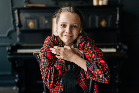Cheerful preteen girl pianist sitting backwards on chair, wearing red plaid shirt, braided hair framing cheerful smile near piano in dark room. Portrait of pretty child posing alone indoors.の写真素材