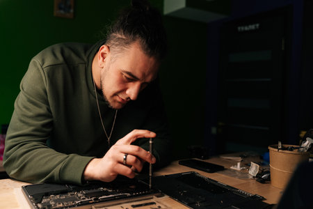 Portrait of technician male carefully using screwdriver to reassemble laptop after performing repairs on motherboard in specialized workshop with dark interior. Process electronic device repair.の写真素材