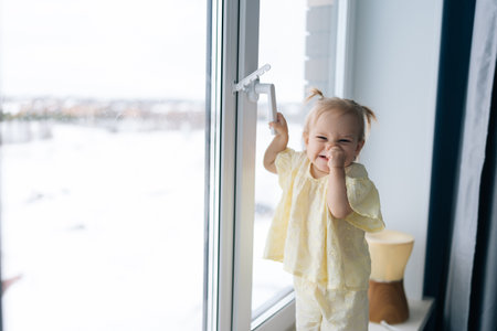 Cheerful toddler girl with two playful ponytails holding window handle while partially covering mouth and beaming with joyful smile. Portrait of cute child wearing yellow dress standing on windowsill.の写真素材