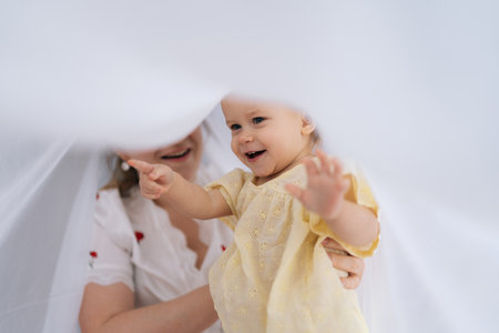 Pretty mother and infant daughter enjoying playful moments together, hiding under soft white blanket, pointing fingers and sharing laughter in comfort of cozy home. Concept of parenting love and care.の写真素材