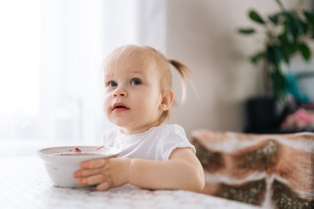 Close-up of cute little child girl with two ponytails eating red beet soup from bowl sitting at table with white tablecloth, enjoying healthy and traditional meal in cozy kitchen room.の写真素材
