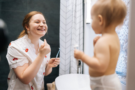 Joyful mother and toddler daughter having fun in bathroom while brushing teeth, toddler girl touching mother nose with finger. Concept of scene curiosity and innocence in bathroom setting.の写真素材