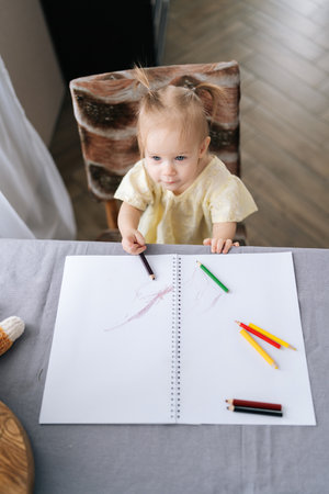 Vertical top view of little girl drawing with colored pencils on paper notebook, sitting at table, expressing creativity and enhancing fine motor skills while enjoying artistic play at home.の写真素材