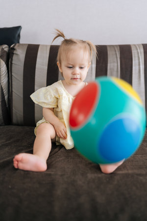 Vertical portrait of toddler girl sitting on comfortable sofa, joyfully playing with large colorful ball with parent, enjoying playful moments in cozy atmosphere at home. Concept of parenting love.の写真素材