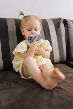 Vertical shot of focused toddler girl engaging with smartphone screen, exploring digital content while sitting comfortably at home, representing early technological interaction alone.の写真素材
