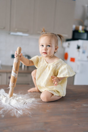 Vertical portrait of cheerful child girl exploring kitchen, playing with wooden rolling pin and pile of flour on table, learning through play and sensory exploration. Concept pleasures of childhoodの写真素材