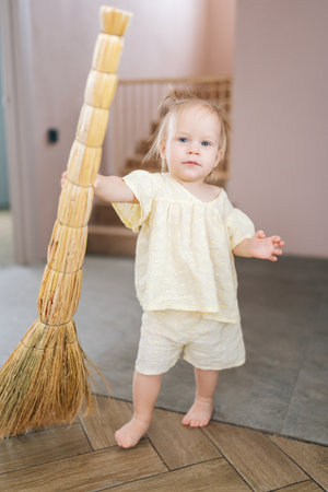 Full length vertical portrait of cute toddler girl standing barefoot on floor holding broom in modern living interior, helping with household chores and learning responsibility from young age.の写真素材