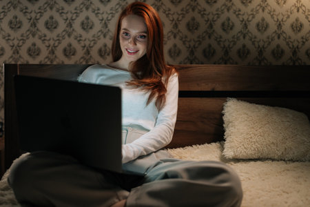 Portrait of happy female freelancer typing on laptop keyboard smiling looking to screen while sitting comfortably in bed, working remotely in cozy bedroom late at night. Concept of modern lifestyle.の写真素材