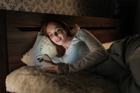Attractive young woman lying in bed at night, using mobile phone smiling looking to screen, enjoying leisure time in cozy bedroom, illuminated by cell screen. Concept of mobile addiction and insomniaの写真素材