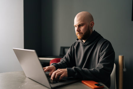 Bald male programmer with beard and hoodie remote working from home sitting at desk and typing on laptop computer, concentrating on tasks in modern living room interior, serious looking to screen.の写真素材